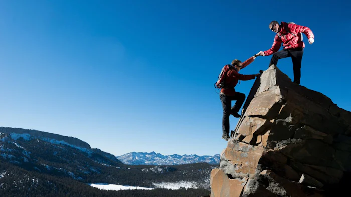 Man helps a climber reach the top of a cliff in cold conditions.
