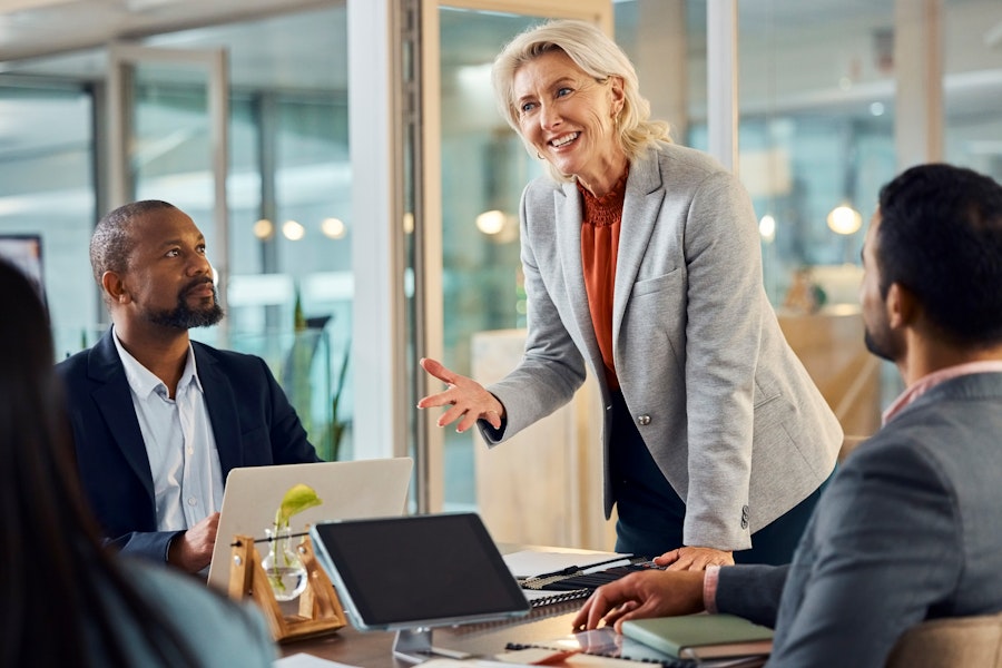 A standing businesswoman talks confidently to seated team members, an example of an effective woman leader.