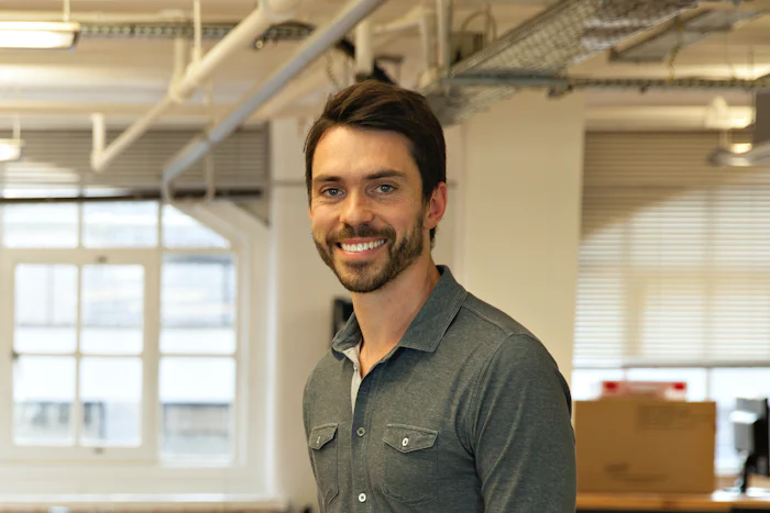 A portrait of Ed Thompson, a white man with dark hair and facial hair, wearing a collared shirt and standing in an office.